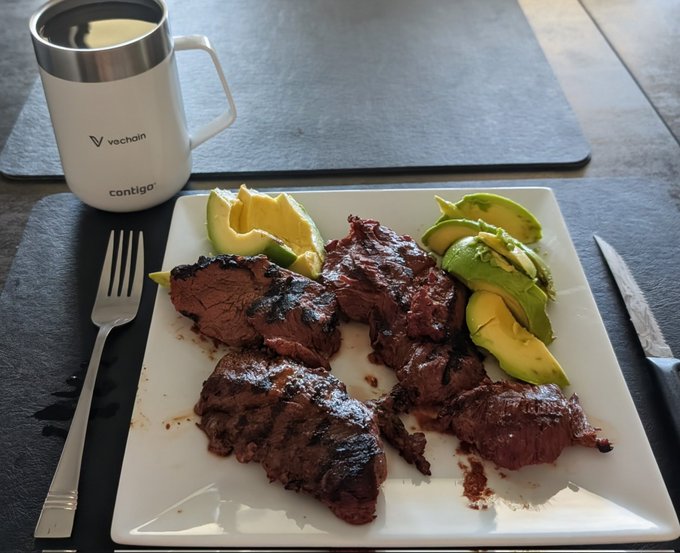 iced coffee, grilled steak with avocado and lime wedges, and a fork rest on a black placemat.