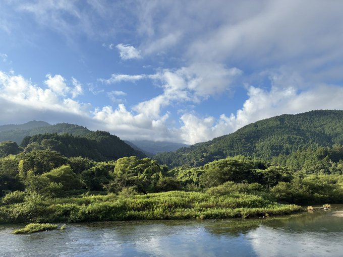 A scenic landscape view of lush green forested mountains rising behind a calm river under a partly cloudy blue sky.
