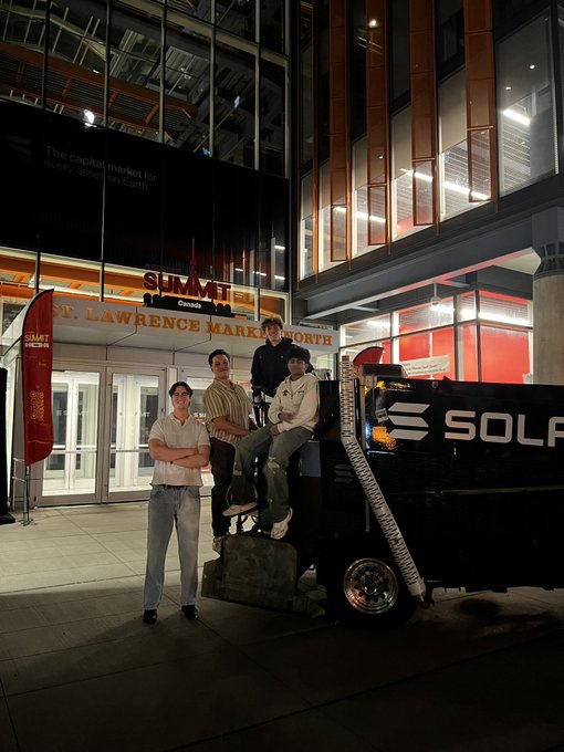Four young people pose with a black vehicle labeled 'SOLAR' parked in front of the Scotiabank Arena and St. Lawrence Market in Toronto at night.