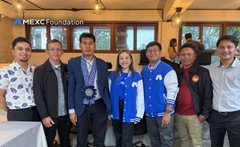 The image features a group of people standing together, posing for a photo. There are at least nine individuals in the picture, all wearing blue jackets with white stripes. The group appears to be from the MEXC Foundation, as indicated by their attire. They seem to be gathered for an event or gathering related to blockchain technology and education.