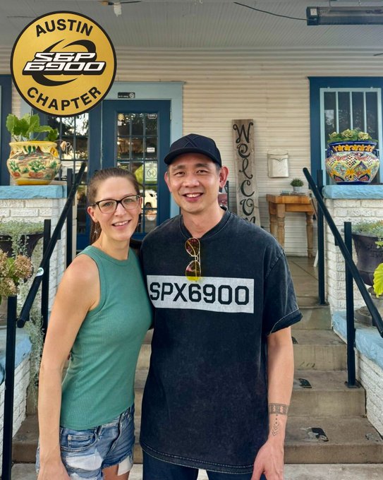 A man and woman posing together on the porch of a light blue house with a 'WELCOME' sign; the man wears a black 'SPX6900' t-shirt and a black cap with sunglasses on his shirt, and an 'Austin SPX6900 Chapter' badge is overlaid in the top-left corner.