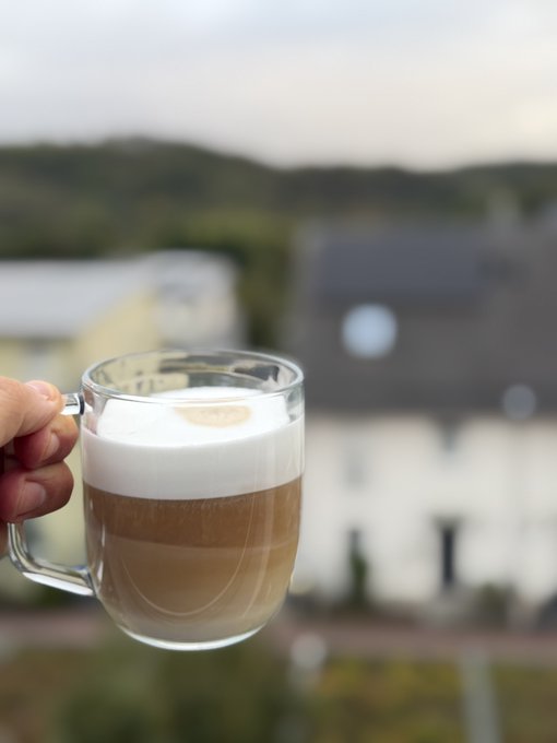 A hand holds up a glass mug of layered latte coffee, with a blurred background of trees and buildings.
