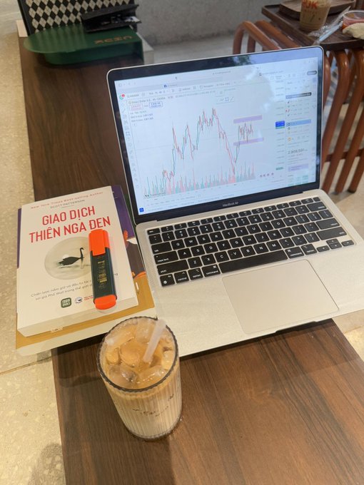 A workspace setup with an open laptop displaying a stock chart, next to a book titled 'GIAO DỊCH THIÊN NGA ĐEN' (Black Swan Trading) by Nassim Nicholas Taleb, an orange highlighter, and a glass of iced coffee on a wooden table.