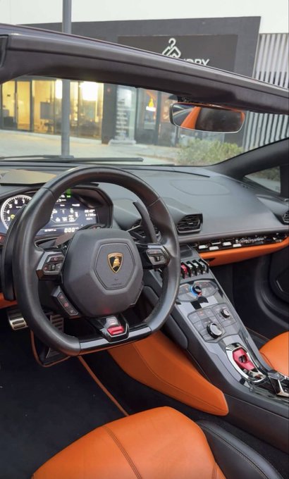 An interior view of a Lamborghini features an orange and black leather cabin, a steering wheel with the Lamborghini shield, and a center console with a red start button cover.