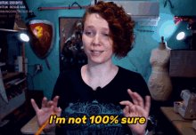 The image shows a woman with red hair sitting in front of a green table and holding up her hands as she speaks into a microphone, possibly discussing her experience with a new domain or app. The background features a sign with the phrase "I'm not 100% sure", which may suggest an ongoing debate or uncertainty related to the subject at hand.