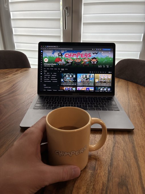 A hand holds a yellow 'CHIMPERS' mug in front of a MacBook Pro displaying the Chimpers Doodles YouTube channel page, sitting on a wooden table near a window.