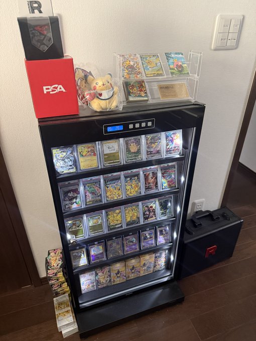 A black display case with LED lighting showcases an extensive collection of graded Pokémon cards housed in PSA slabs, alongside a red PSA storage box and a Dedenne plushie on top. The setup reflects the collector's concern about humidity control—essential for protecting valuable collections that have reached five to six figures in value.