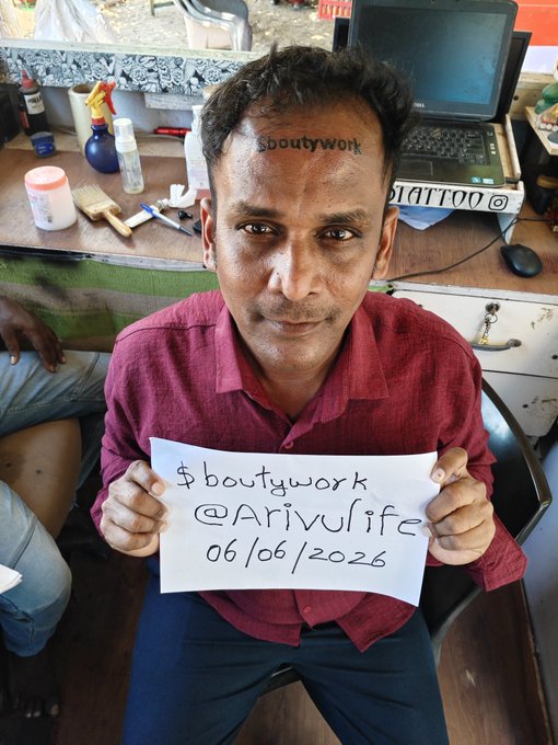 The image features an Indian man sitting down with a sign that says "bouttywork" written on his forehead, which has been tattooed by someone else. The man is holding up this sign for the camera, possibly to show off his new tattoo or to share it with others. The photo also includes other people in the background and a laptop computer, suggesting that they might be in an office setting.