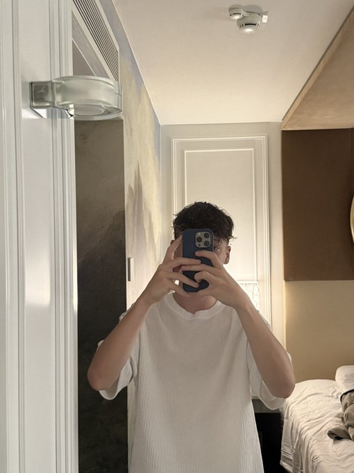 A person taking a mirror selfie in a bedroom, covering their face with a phone. The cozy, modern room with warm lighting and minimal decor complements the casual "GM" (Good Morning) greeting shared in the post.