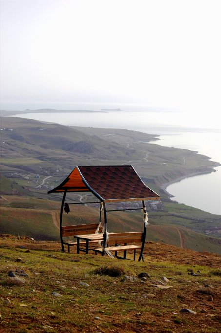 Scenic landscape photograph of a wooden shelter with a curved shingled roof and bench seating, situated on a grassy hilltop overlooking a vast coastal inlet with islands visible in the misty distance.