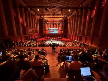 An audience seated in a grand, dimly lit auditorium watching a presentation on stage.