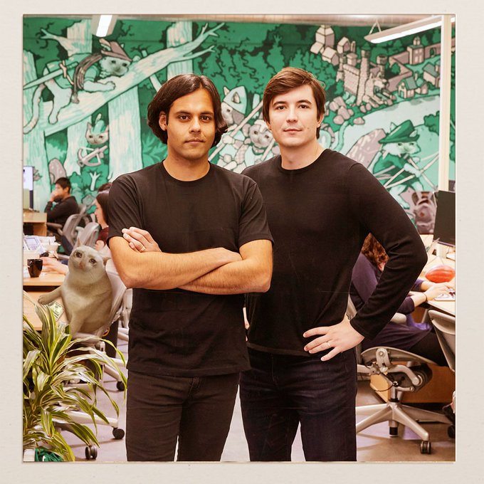 Two casually-dressed young men in black shirts pose confidently with crossed arms in an office setting, with a whimsical cartoon mural and an unusually realistic cat (or cat mascot) in the background — the "lock in mf" post suggesting they've both entered intense focus mode, matching their serious, determined expressions.