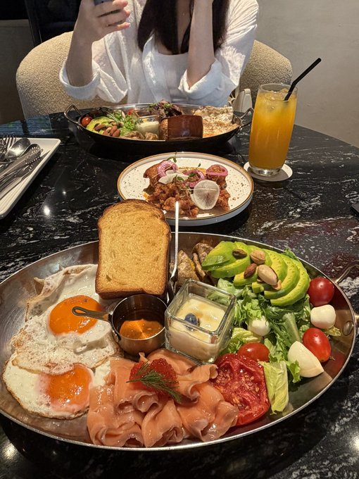 A table set with a large plate of food including fried eggs, toast, smoked salmon, avocado, and salad, alongside orange juice.