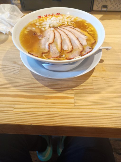 A bowl of ramen or similar noodle soup with sliced pork/chashu served in a white bowl with broth and green onions, placed on a wooden table.