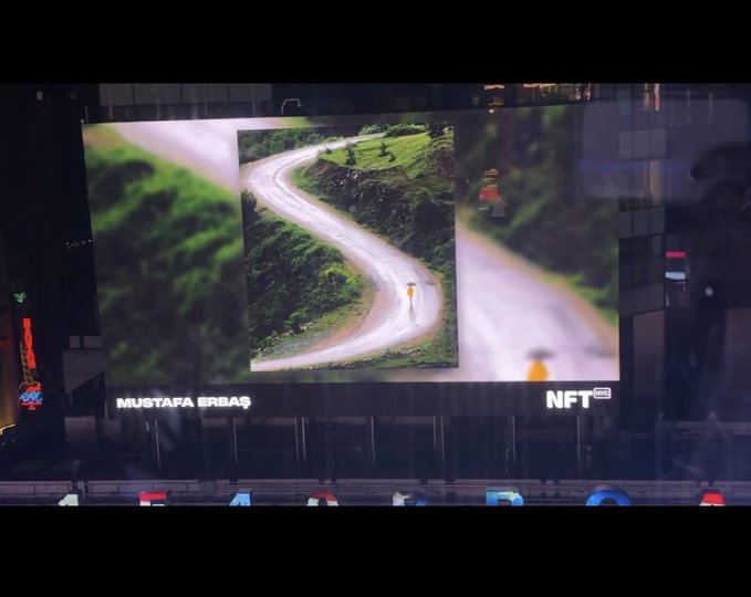 A large billboard displaying an aerial image of a winding road with text 'MUSTAFA ERBAŞ' and 'NFT.'
