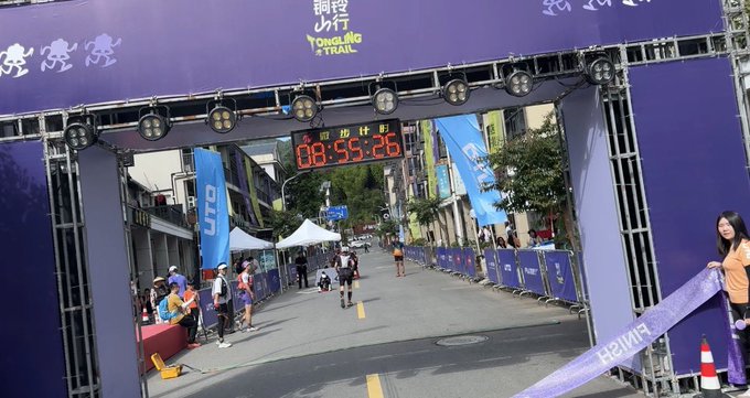 The image shows a runner crossing the finish line of a race, with a purple archway or gate marking their accomplishment. A woman stands next to the runner, possibly a race official or participant, as they both look at the clock on the wall displaying the time remaining in the race. The scene captures the excitement and sense of achievement that comes from completing a challenging event like a marathon.