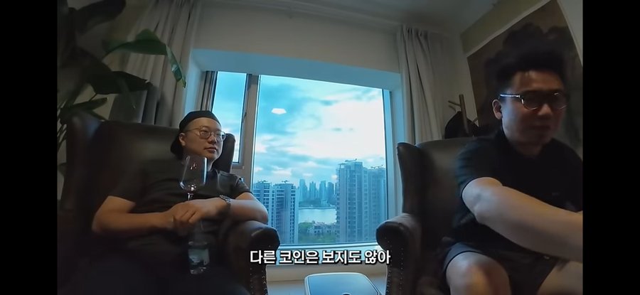 Two men sit in armchairs by a window overlooking a city skyline, with Korean subtitles reading "다른 코인은 보지도 않아" (I don't even look at other coins), reinforcing the post's message to only invest in Bitcoin and ignore altcoins.