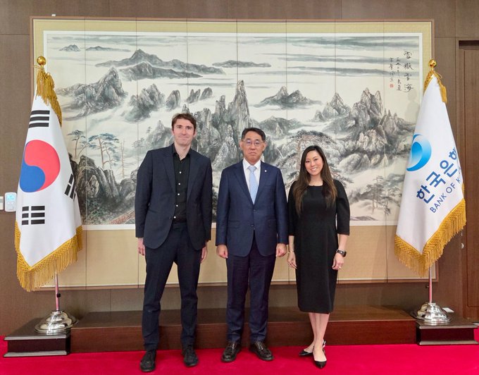 A formal photo of three people (two men in suits and one woman in a black dress) standing between a South Korean flag and a Bank of Korea flag, with a traditional Korean landscape painting as the backdrop.