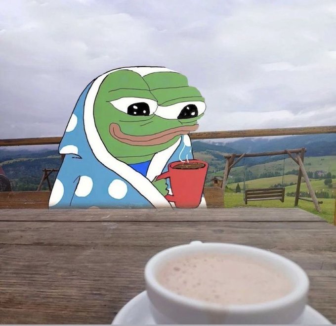 A Pepe the Frog meme cartoon character wearing a blue polka-dot robe holds a red mug while sitting at a wooden table overlooking a mountain landscape, with a coffee cup in the foreground.