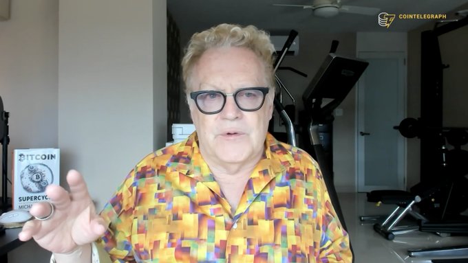 A man in a colorful pixel-patterned shirt speaks on a Cointelegraph video interview, with a "Bitcoin Supercycle" book visible in the background, discussing Bitcoin's market outlook and its appeal as a long-term investment.
