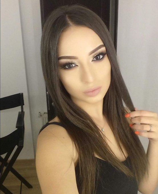 A selfie of a woman with long brown hair, heavy eye makeup, orange nails, and a ring, wearing a black top.