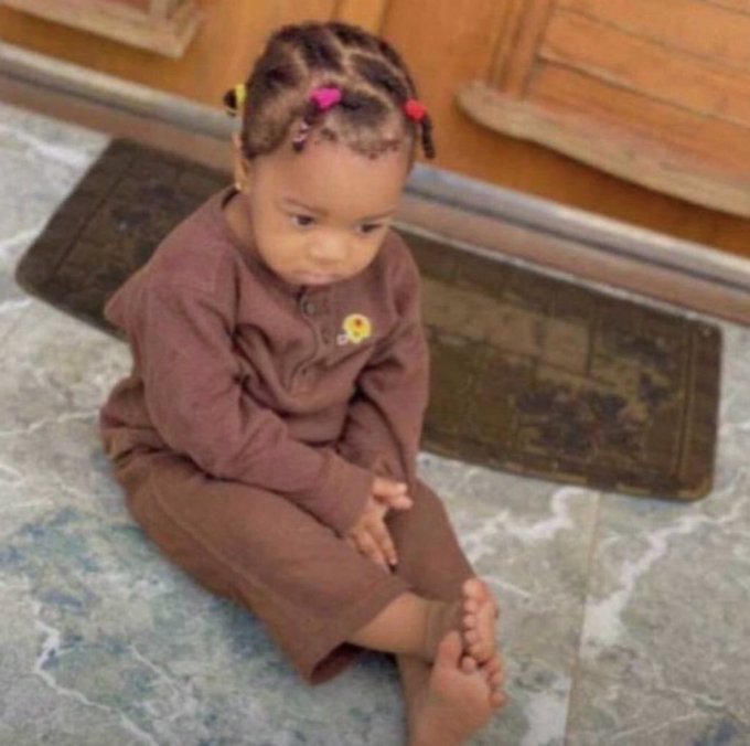 A young child with braided hair sits on a tiled floor, wearing a brown outfit and looking down with a contemplative or sad expression.