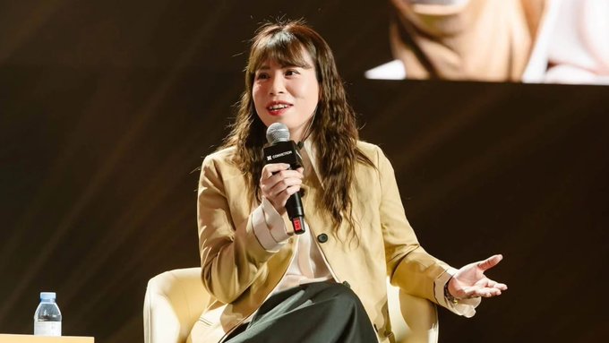 A woman with long wavy hair wearing a tan blazer speaks into a microphone while seated on stage at an event.