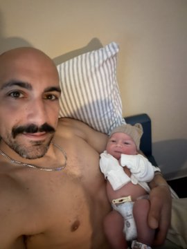 The image features a man holding a baby while lying down on a bed. The man has a beard and mustache, and he appears to be balding. He is wearing a necklace around his neck. The baby is dressed in white clothing and is being held by the man's arms. In the background of the photo, there are two pillows visible - one located behind the man on the left side and another positioned above him on the right side.