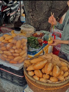 A street food vendor scene showing woven baskets and plastic bags filled with small bread rolls, along with containers of dipping sauces and vegetables, with two people's hands preparing food with chopsticks.