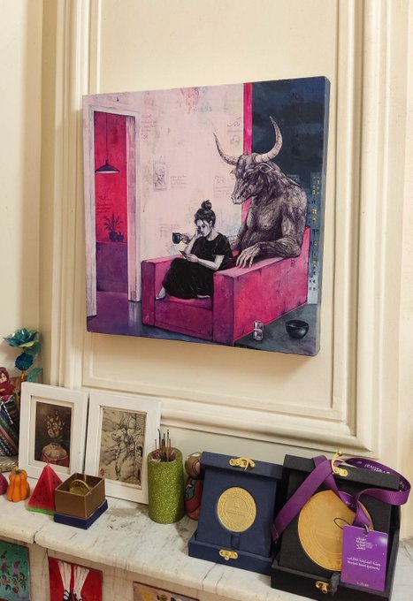 A pink and black painting of a bull-headed figure and a woman sitting together on a couch hangs on a cream wall above a mantel displaying framed photos, decorative objects, and packaged chocolate coins.