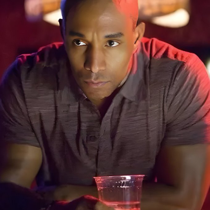 A young man with a serious expression sitting at a bar, lit by red lighting, with a cocktail glass in front of him.