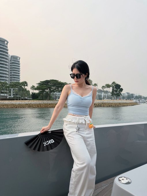 A young woman wearing sunglasses, a light blue top, and white pants, standing by a waterfront while holding a black folding fan with 'EXO' printed on it.