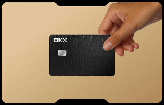 A hand holding a black OKX-branded credit/debit card against a tan background.