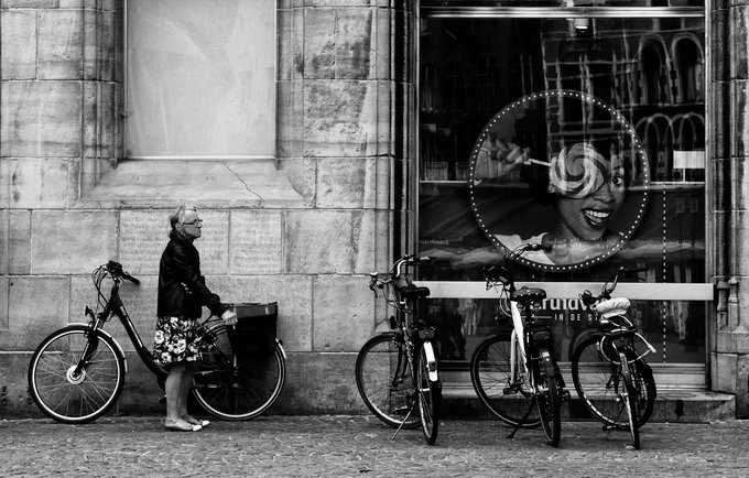 A black-and-white street photograph showing an older woman with a bicycle standing on a sidewalk next to a building with large display windows, where a circular sign featuring a smiling woman eating a lollipop is visible; multiple other bicycles are parked nearby.