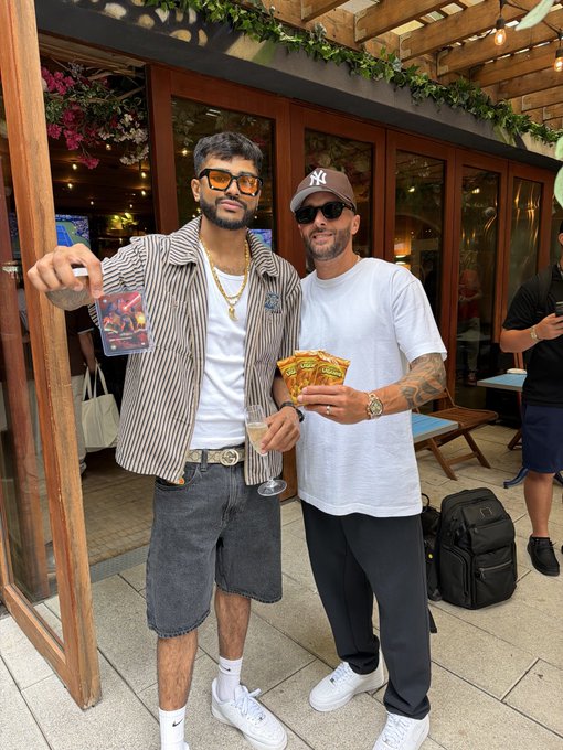 Two men pose together outside a venue with greenery; one wears a striped jacket, denim shorts, and sunglasses while holding a colorful card and a drink, and the other wears a white t-shirt, NY cap, and holds yellow tickets.