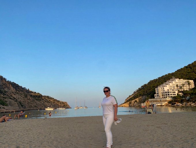 A woman in white clothing and sunglasses stands on a sandy beach with sailboats in the water and hillside buildings in the distance.