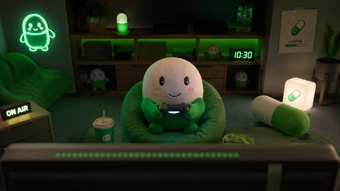 A cute pill-shaped mascot relaxes on a green beanbag while playing video games in a cozy, neon-green lit gaming setup, with a digital clock reading "10:30" and an "ON AIR" sign visible. The image playfully ties to the post's caption, showing the mascot taking a relaxed break while the community is called upon to send @CupseyToken to 500M market cap.