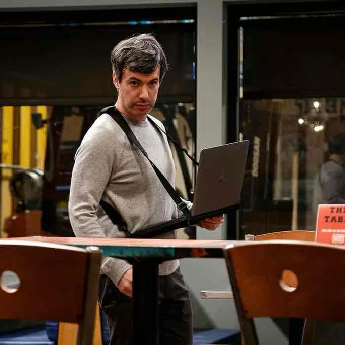 A man with graying hair wearing a gray sweater and a harness holds a laptop while standing between chairs in what appears to be a cafe or restaurant.