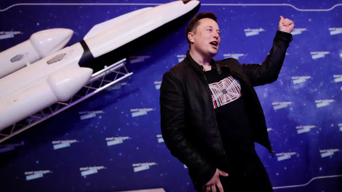 Elon Musk speaking at an event with a SpaceX rocket model in the background, gesturing with his arm raised.