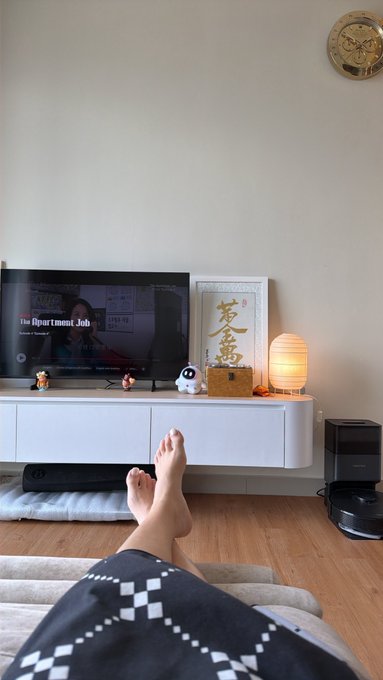 A person relaxes barefoot in their living room, watching "The Apartment Job" on TV—likely a Korean drama as part of a Binge Watch Challenge—while a "Lucky" (君盈) Chinese calligraphy print and warm lantern create a cozy, unwind-from-work atmosphere.