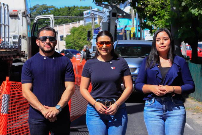 Three people stand together on a sunny street in El Salvador, with the man on the left and the two women on the right all wearing coordinated navy blue tops. The image reflects the post's theme of national progress under President Nayib Bukele, showcasing citizens who appear to be part of a media or reporting team documenting the country's development.