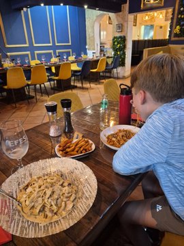 A young boy sits at a restaurant table enjoying a plate of fettuccine pasta, surrounded by other dishes and a red water bottle. The image reflects the post's message about prioritizing family moments and good food over fleeting trends.