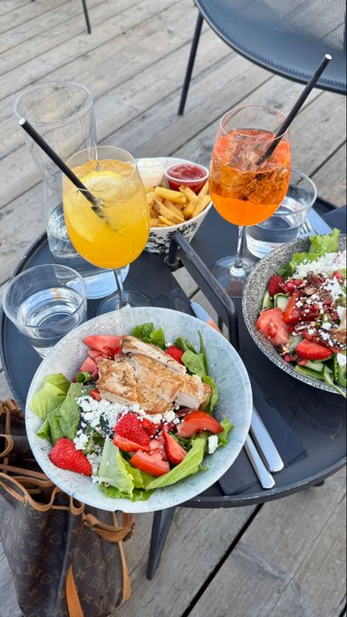 An outdoor dining scene featuring two bowls of salad with grilled chicken and strawberries, french fries, and two glasses of orange-colored beverages with straws on a dark table.