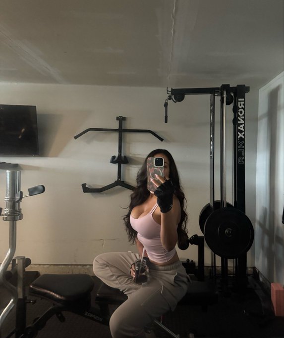 A woman takes a mirror selfie in a home gym, wearing a pink top and beige pants, with a RIXMAXORI cable machine visible behind her.