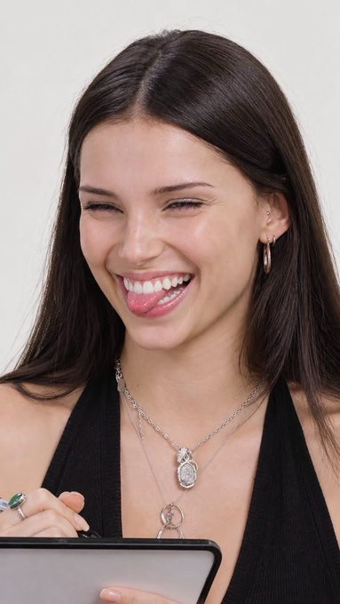 A smiling woman with long dark hair, wearing layered silver necklaces and a black halter top, laughs joyfully while looking down at what appears to be a tablet or device. Her wide, delighted expression perfectly matches the post's humor—capturing the ironic excitement of reacting positively to unexpected news, like the thrill of jumping into crypto despite financial uncertainty.