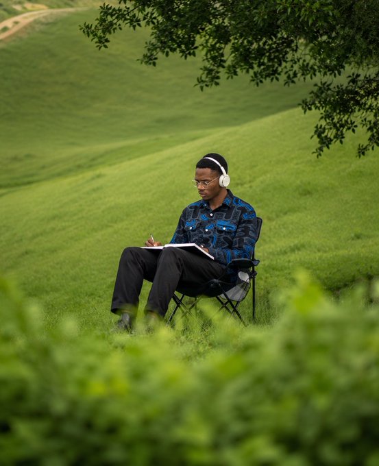 A young man wearing headphones, glasses, and a patterned blue shirt sits on a folding chair in a lush green field, writing in a notebook.