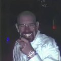 A dark, low-quality photo of a bald man in a white shirt holding his hand near his face in a dimly lit setting.