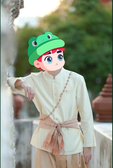 A young person wears traditional light-colored attire with a cross-body sash, with a cartoon frog hat emoji edited onto their face.