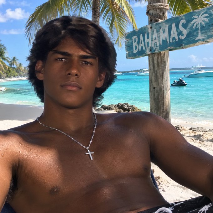 A shirtless young man with dark curly hair and a silver cross necklace posing at a tropical beach location, with a wooden 'BAHAMAS' sign, palm trees, turquoise water, and boats visible in the background.