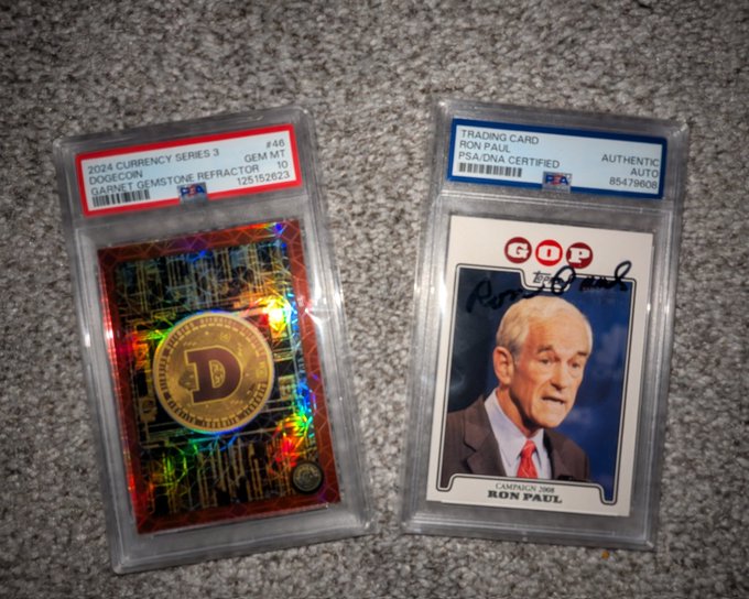 The image shows two trading cards placed side by side on a gray carpeted floor. The cards are from the 2016 currency series, featuring a man's face with the words "DOGE" written on them. There are also other cards displayed in the background, including sports and Pokemon cards.
